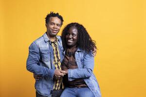 Romantic african american life partners embracing each other on camera, smiling and feeling cheerful against yellow background. Joyful pleased husband and wife hugging with affection. photo