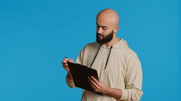 Middle eastern adult creating a checklist on clipboard papers, being focused on trying to plan tasks on an organizer in studio. Modern person looking at files and listing things to do. Camera 1. photo