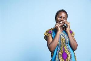 Excited woman using her mobile device to take calls, chuckling while she is engaging in a remote discussion with her friends against blue backdrop. In studio, african american person communicates. photo