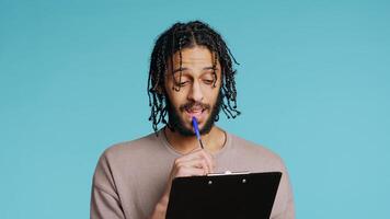 Middle Eastern photographer holding clipboard in hand, completing shoot list for upcoming photography work. Creative man thinking of ideas, writing on paper using pen, studio background, camera B photo