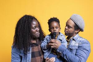 African american couple making their toddler sing in front of the camera, using microphone and having fun together. Loving parents listening to their child singing in studio, talented young boy. photo