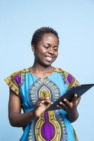 Young person explores social media posts on a blue backdrop using portable device. In front of the camera, african american joyful woman uses a tablet to surf the web for updates. photo