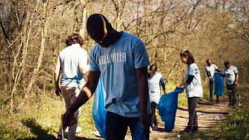 Eco friendly activist grabbing garbage and plastic waste from a forest area, cleaning up the woods and collecting rubbish in a trash bag. Man volunteer dealing with waste management. Camera B. photo