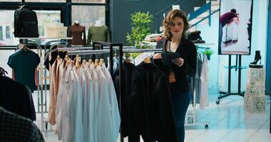 Woman employee examining racks of clothes for stock inventory, using tablet to check all products. Retail store assistant checking discount list on device, ensuring commercial tags. Camera B. photo