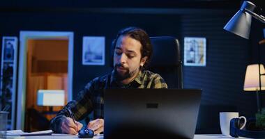 Entrepreneur working at home office desk on laptop, browsing the internet and writing in notebook. Caucasian man using pen, notepad and digital device while teleworking, camera B photo