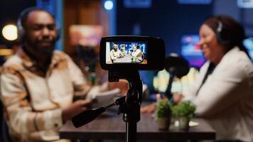 Focus on professional camera used to record radio show presenter live conversation with man in personal studio. Close up on production gear with host and guest talking in blurry background photo