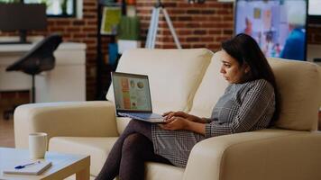 Remote employee sitting on couch in apartment, optimizing market performance charts while working from home. Woman checking online business analytics statistical data metrics, camera B photo