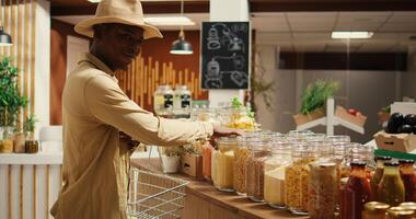 African american client shopping for pasta or grains in jars, looking at variety of bulk products stored in reusable containers. Buyer supporting eco friendly business, nonpolluting goods. Camera 2. photo