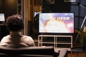 Man looking at game over screen of science fiction videogame on smart TV. African american gamer receiving defeat message after failing to beat level, watching statistics roll on display photo