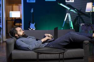 Freelancer lounged on couch working from home in apartment solving business related tasks on laptop. Remote employee laying down in neon lit living room, sending emails on notebook photo
