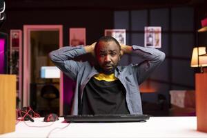 Shocked gamer putting hands on head after seeing game over screen, losing singleplayer videogame on gaming computer at home. Portrait of african american man being upset after being defeated photo