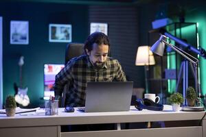 Working indoors, a tech-savvy man sits at a table, easily demonstrating multitasking skills. Bearded guy preparing to use his laptop for research and checking of emails. photo