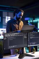 IT professional working from RGB lit home using laptop, testing and deploying programs and systems. Software engineer checking code displayed on computer monitors, standing in apartment photo