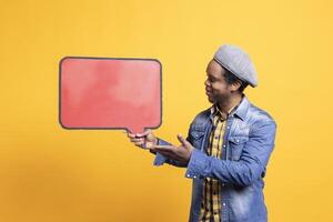 Cool mature person showing speech bubble red icon on camera, holding an empty billboard copyspace sign to create a web commercial ad. African american guy shows cardboard cut out. photo