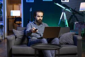 Portrait of cord cutter bored at home using TV to watch films on streaming services and typing on laptop. Man in living room mindlessly consuming entertainment with notebook in lap photo