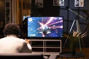 African american man in home theatre using cloud gaming service to play demanding science fiction videogame on TV. Gamer enjoying high quality graphics, streaming game over the internet photo
