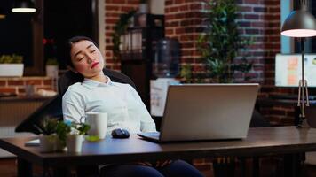 Sleepy employee asleep on computer desk at night in brick wall office, suddenly waking up. Tired businesswoman looking at watch, annoyed by time passing too slow while working overnight, camera B photo