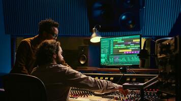 Musician and producer work on soundboard pads at professional studio station. Sound designer uses digital mixer in music production, operating on sliders and key buttons at control desk. Camera B. photo