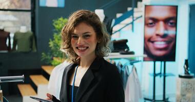 Smiling retail clerk enjoys working in a fashion boutique, verifying stock on hangers and racks. Employee using tablet to check inventory items off of list, arranging clothes on discount. Camera B. photo