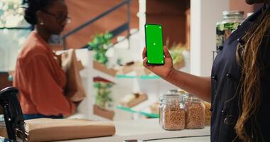 Store vendor holding phone with greenscreen display, working at cash register to serve customers. African american seller using isolated mockup template on smartphone screen. Tripod shot. photo