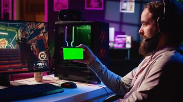 Man watching tutorial on mockup phone while playing first person shooter videogame with gun shooting laser bullets. Gamer learning to play game by looking at online guide on chroma key device photo