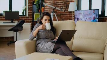 Teleworker lying on couch, drinking coffee while remotely performing tasks in personal office. Freelancer feeling stressed, enjoying hot beverage to relax while inputting data on laptop, camera A photo