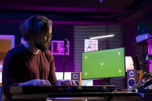 Music producer composing a melody on midi controller keyboard and watching online lessons on computer with greenscreen. Songwriter playing electronic piano in home studio, producing tunes. photo