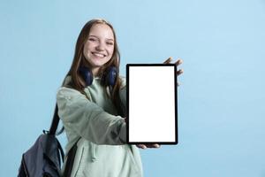 Jolly young girl presenting tablet with isolated screen display, doing advertisement, smiling, studio background. Merry teenager creating ad promotion, showcasing mockup device photo