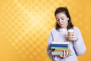 Woman holding pile of books, enjoying cup of coffee in the morning. Tired person with stack of novels in arms, drinking caffeinated beverage, isolated over studio background photo