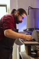 Carpenter in woodworking shop wearing protection goggles while using disc sander to avoid injury. Cabinetmaker in joinery using professional grinding machine with safety gear on to prevent accidents photo