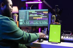 Sound designer producing new soundtracks with digital software, having a tablet with greenscreen on desk. Young talented artist mixing and mastering his audio files, editing in post production. photo