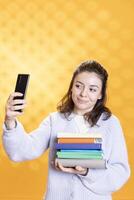 Smiling woman holding pile of books, enjoying reading hobby, taking selfies with cellphone. Cheerful lady with stack of novels in arms doing pictures with mobile phone, studio background photo