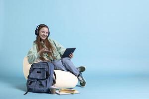 Smiling teenager wearing headphones having fun, using tablet, being entertained. Jolly person relaxing by listening music, picking songs in playlist on device, studio background, photo
