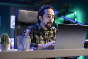 Bearded man in checkered shirt entering data on modern laptop while seating on chair indoors. Male entrepreneur reviewing annual financial report using electronic device. photo