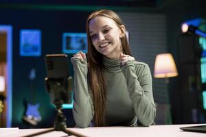 Happy content creator records introductory with smartphone, inviting viewers to subscribe to online channel. Media star uses phone to film herself connecting with watching audience photo