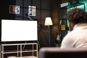 BIPOC man watching subscription based football competition live on isolated screen smart TV. Homeowner on couch in front of mockup television set display entertained by soccer match photo