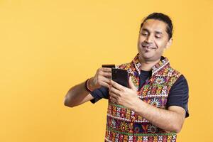 Smiling man looking at debit card to read banking account, feeling happy to do online purchases, studio background. Cheerful man does internet shopping using cellphone, having fun photo