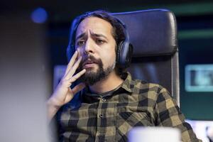 Closeup of male college student taking break from studying by watching a sad film on his laptop. Bearded man with wireless headset viewing a movie on his digital device with sorrowful expression. photo