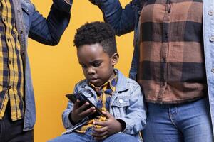African american small toddler looking at something on smartphone, parents keeping him busy against yellow background. Adorable little baby boy using a mobile phone in studio. photo