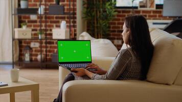 Self employed woman staying at home using green screen laptop to do various job tasks from couch. Remote working indian person typing data on chroma key notebook keyboard photo