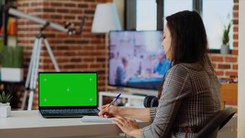 Teleworker staying at home using green screen laptop to do various job tasks at wooden desk. Remote working employee typing data on chroma key notebook keyboard and writing on notepad photo