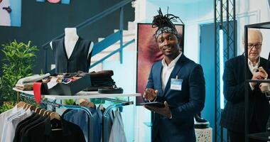 African american worker managing clothes stock on hangers, working on inventory using tablet in shopping center. Male store assistant in a suit preparing fashion collection on display. Camera B. photo