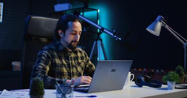 Smiling man sitting at home office desk and typing on laptop keyboard, solving tasks. Happy remote worker using notebook to send emails to business partners from living room, camera B photo