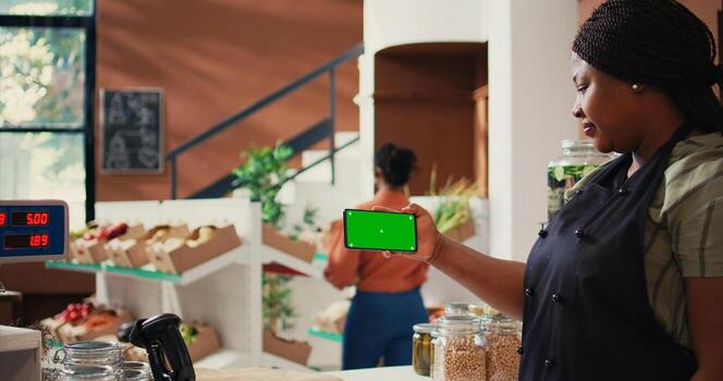 Storekeeper holds mobile phone with greenscreen display while she stands at the checkout counter. African american merchant showing isolated chromakey layout on mobile phone app. Tripod shot. photo