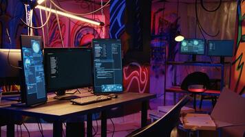 Computer screens showing malware development code in rundown underground hideout with graffiti painted on walls. PC system equipment used for cybercriminal activity in neon lit place photo