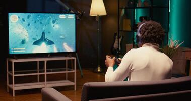 BIPOC man plays SF game with overlay HUD showing health bars and minimap on TV in home theatre. Player enjoying high quality ray tracing graphics on console system, jib down shot photo
