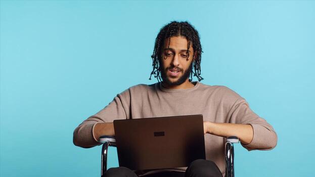 Man in wheelchair chatting with friend over internet videocall using laptop application. BIPOC person with paraplegia chatting with mate in videoconference online meeting, studio background, camera A photo