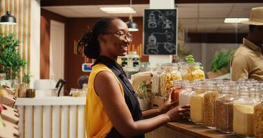 African american vendor arranging bulk products at local store, promoting zero waste concept for sustainable lifestyle. Merchant smiling and preparing to welcome clients at bio shop. Camera 2. photo