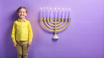 A boy in a yellow shirt and yellow pants stands with a candle in the form of a menorah on a purple background. photo