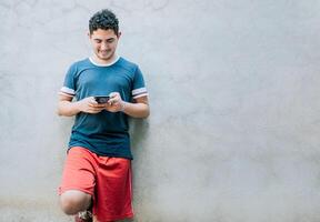 Happy guy leaning on a wall using smartphone. Smiling young man leaning on a wall sending a text message photo